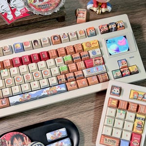 May include: A custom white keyboard with character-themed keycaps, including a small screen. The keycaps feature various cartoon characters and designs. Additional accessories include a matching number pad, coasters, and a figurine.