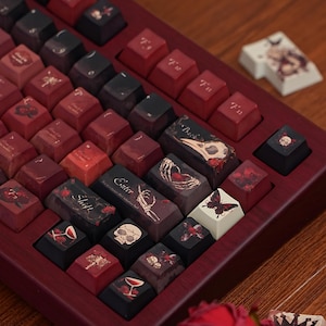 Puede incluir: Un teclado mecánico personalizado con teclas en tonos rojos, negros y burdeos. Las teclas presentan diseños de inspiración gótica, incluyendo calaveras, mariposas y motivos florales. El teclado está enmarcado en un marco de madera oscura.