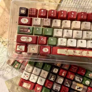 Könnte beinhalten: Eine Sammlung dekorativer Tastaturkappen in einem transparenten Kunststoffbehälter. Die Tastenkappen weisen verschiedene Designs auf, darunter florale Muster, geometrische Formen und Illustrationen. Die Farbpalette umfasst Rot, Grün, Weiß und Schwarz.