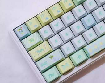 May include: A pastel-colored keyboard with white, yellow, and blue keycaps. The keys feature letters, numbers, and symbols, with some keys displaying decorative illustrations and text such as "Tab", "Caps", and "Shift". The keyboard has a white frame.