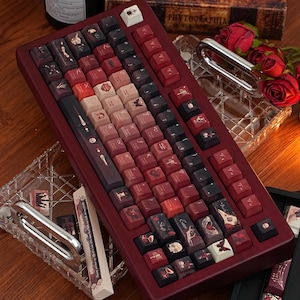 May include: A burgundy mechanical keyboard with custom keycaps. The keycaps feature illustrations in black, red, and gold, including skulls and butterflies. The keyboard has a wooden frame. Decorative items such as roses and a glass dish are in the background.