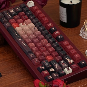May include: A custom mechanical keyboard with a dark red wooden frame and keycaps in shades of red, black, and cream. The keycaps feature various illustrations, including skulls, butterflies, and floral designs. A black candle sits in the background.
