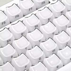 May include: Close-up of a white keyboard with rounded keycaps. The keys are arranged in rows and columns, with letters, numbers, and symbols visible. The keycaps have a glossy finish and a dark border.