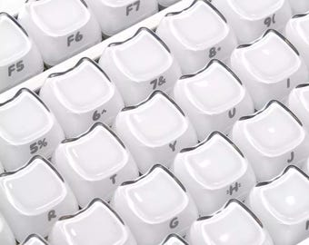 May include: Close-up of a white keyboard with rounded keycaps. The keys are arranged in rows and columns, with letters, numbers, and symbols visible. The keycaps have a glossy finish and a dark border.
