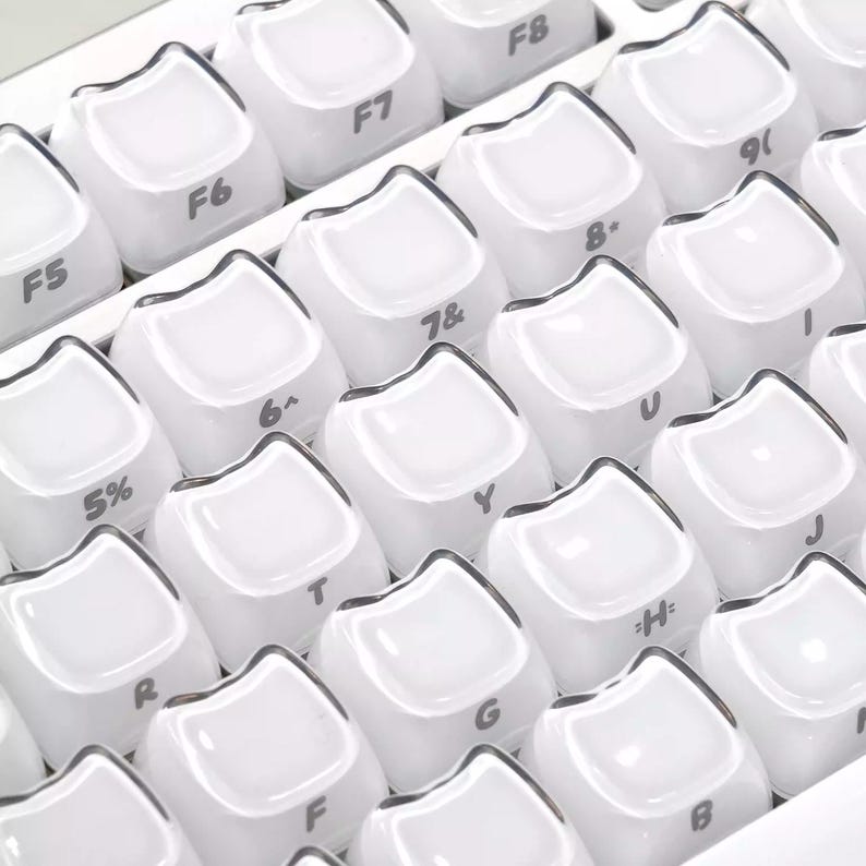 May include: Close-up of a white keyboard with rounded keycaps. The keys are arranged in rows and columns, with letters, numbers, and symbols visible. The keycaps have a glossy finish and a dark border.