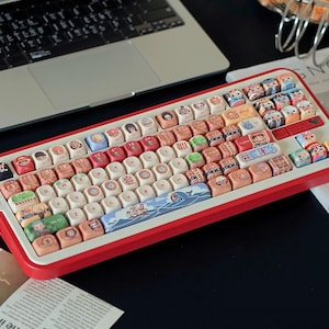 May include: A red and white mechanical keyboard with custom keycaps featuring various cartoon illustrations. The keyboard is positioned next to a laptop and a magazine. The keycaps have a variety of colors and designs.