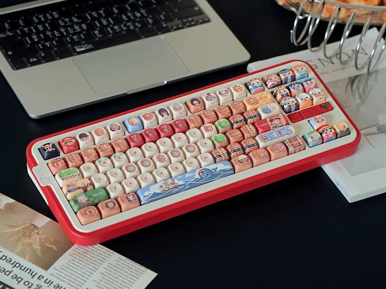 May include: A red and white mechanical keyboard with custom keycaps featuring various cartoon illustrations. The keyboard is positioned next to a laptop and a magazine. The keycaps have a variety of colors and designs.