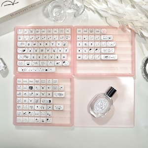 May include: A pink keyboard case with white keycaps featuring cartoon animal designs. The case is open, revealing the keyboard layout. A perfume bottle sits in the lower right corner. The text "Rayon de soleil" is visible on a book in the background.
