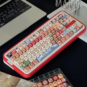 May include: A red and white mechanical keyboard with custom keycaps. The keycaps feature various colorful illustrations and designs. A laptop and a tray of extra keycaps are also visible.