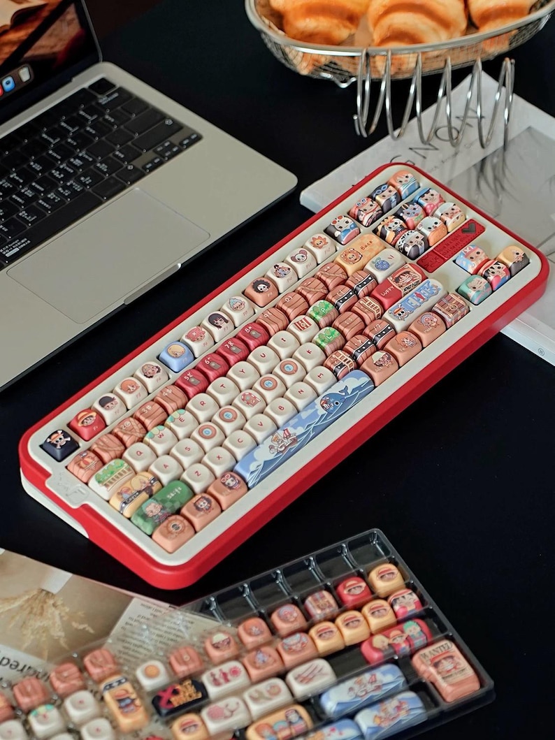 May include: A red and white mechanical keyboard with custom keycaps. The keycaps feature various colorful illustrations and designs. A laptop and a tray of extra keycaps are also visible.