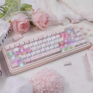 May include: A pastel pink mechanical keyboard with white keycaps and a clear cover. The keyboard has a light pink frame and features colorful LED backlighting. Decorative elements include pink roses and small heart-shaped accents.