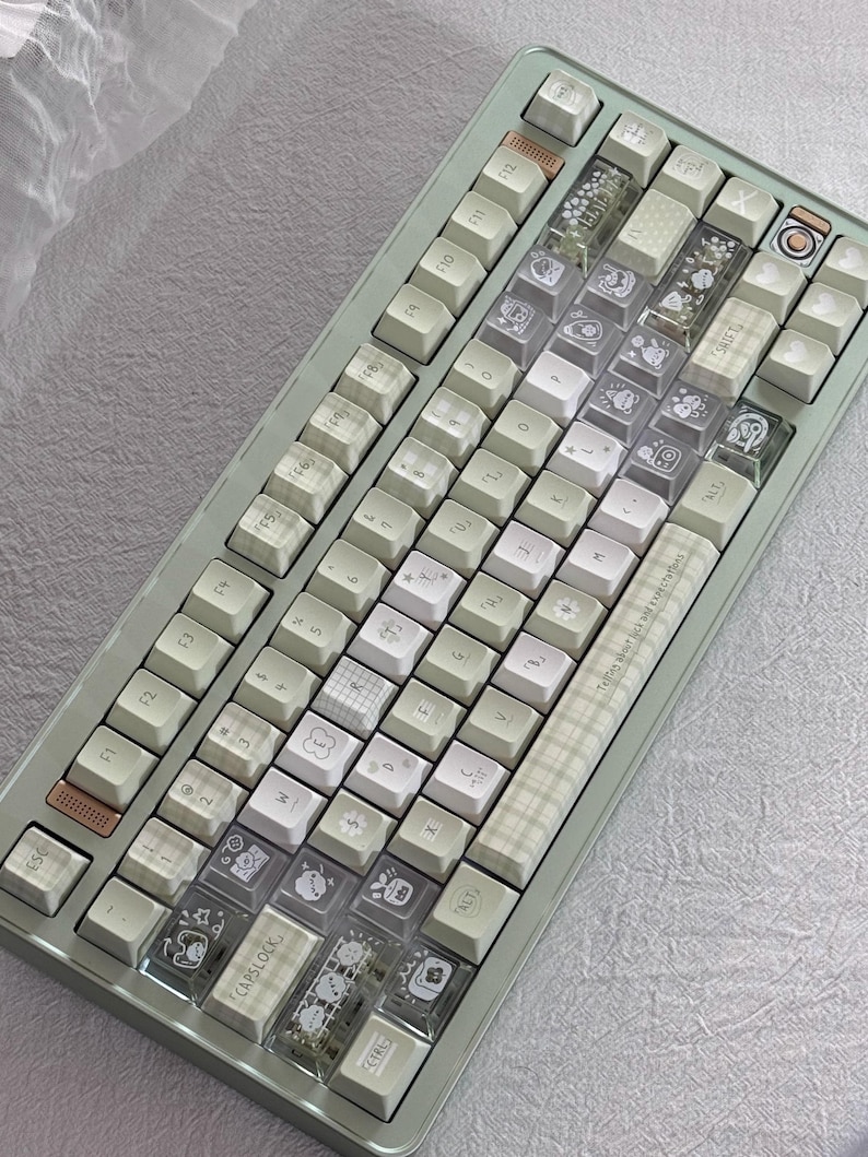 Puede incluir: Un teclado mec&aacute;nico verde menta con teclas blancas y transparentes. Algunas teclas tienen ilustraciones y patrones lindos. El teclado tiene una mezcla de teclas est&aacute;ndar y personalizadas, con el texto "Telling about lack am expectations".