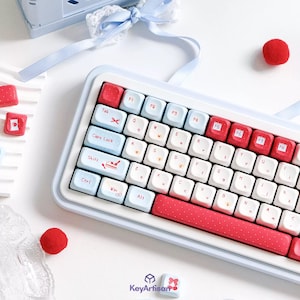 May include: A mechanical keyboard featuring a light blue frame and a mix of white, light blue, and red keycaps. The red keys have white polka dots. Specific keys display icons including scissors, a slice of cake, and a bow. The text KeyArtisan is visible.