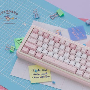 Puede incluir: Un teclado mecánico rosa y blanco con un marco rosa claro. Las teclas son una mezcla de blanco y rosa, algunas con patrones decorativos. El teclado está sobre una esterilla de corte azul con notas y clips.