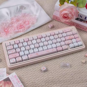 May include: A pastel pink and white mechanical keyboard with custom keycaps. The keycaps feature a mix of pink, white, and light blue colors with decorative illustrations. The keyboard is surrounded by decorative items, including a rose and books.