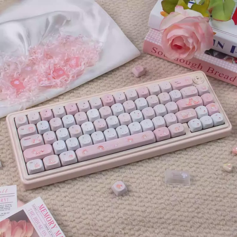 May include: A pastel pink and white mechanical keyboard with custom keycaps. The keycaps feature a mix of pink, white, and light blue colors with decorative illustrations. The keyboard is surrounded by decorative items, including a rose and books.