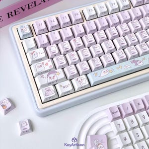 May include: A light purple mechanical keyboard with ghost-themed keycaps. The keyboard has a light blue frame and includes extra keycaps. The design is whimsical and playful, suitable for gamers and keyboard enthusiasts.