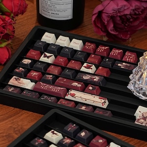 May include: Custom keyboard keycaps in a black tray, featuring designs like butterflies, musical notes, and floral patterns. The keycaps are in white, black, and red hues. A black candle and roses are visible in the background, adding to the aesthetic.