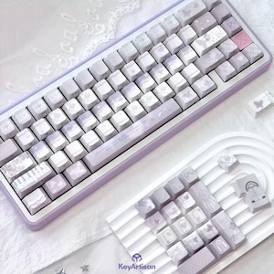Op de afbeelding: Een lavendelkleurig en witte mechanische toetsenbord met aangepaste keycaps. De keycaps hebben bloemen- en wolkenontwerpen, met een paar roze accenttoetsen. Het toetsenbord heeft een bijpassend lavendelkleurig frame en wordt getoond met extra keycaps op een witte tray.
