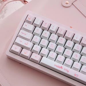 Op de afbeelding: Een roze en wit toetsenbord met Hello Kitty-branding. Het toetsenbord heeft witte toetsen met roze letters en illustraties. Het toetsenbord ligt op een roze ondergrond en de tekst "Hello Kitty" is zichtbaar op het toetsenbord.