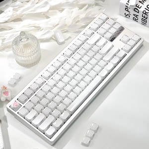 May include: A white mechanical keyboard with white keycaps. The keyboard has a silver frame and is on a white surface. Additional keycaps are scattered around the keyboard. The keyboard is a modern computer accessory.