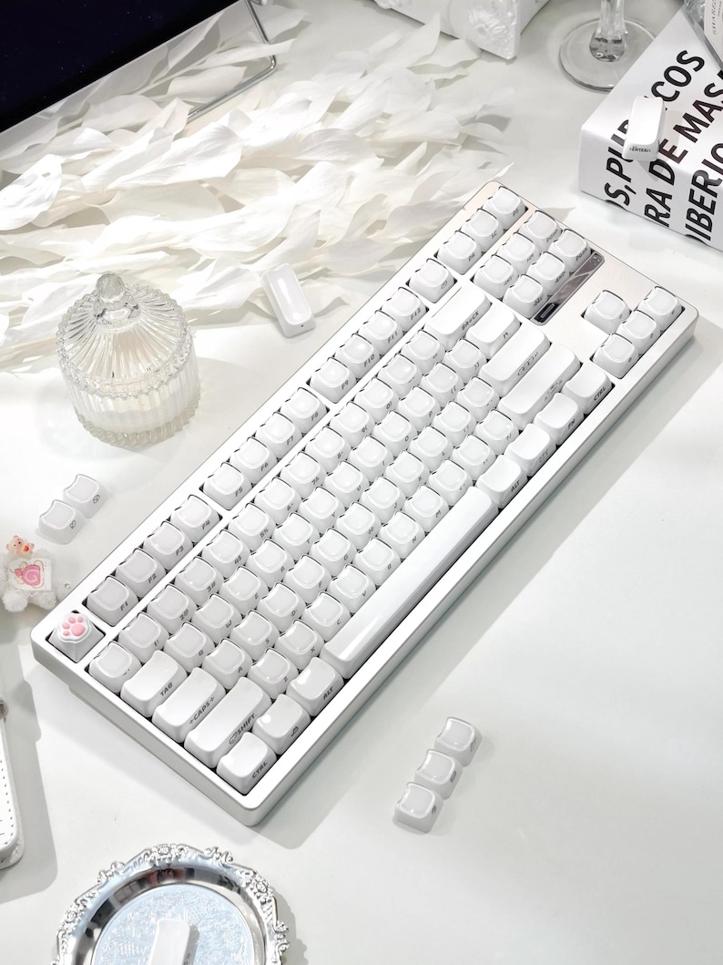 May include: A white mechanical keyboard with white keycaps. The keyboard has a silver frame and is on a white surface. Additional keycaps are scattered around the keyboard. The keyboard is a modern computer accessory.