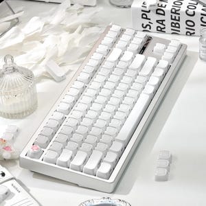 May include: A white mechanical keyboard with white keycaps. The keyboard has a rectangular shape and a minimalist design. The keycaps are arranged in a standard layout. The keyboard is on a white surface.