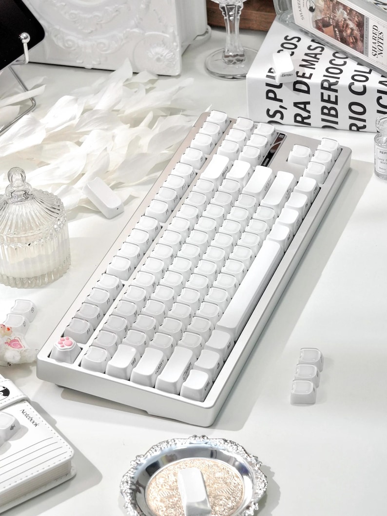 May include: A white mechanical keyboard with white keycaps. The keyboard has a rectangular shape and a minimalist design. The keycaps are arranged in a standard layout. The keyboard is on a white surface.