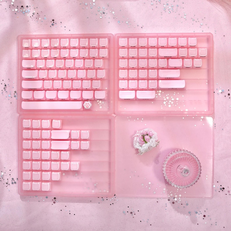 May include: A pink mechanical keyboard with white keycaps, a paw print key, and a clear pink case. The image also shows a pink decorative tray with a floral arrangement and a small, pink, decorative glass dish.