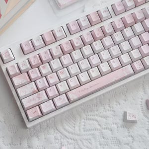 May include: A white mechanical keyboard with pink and white keycaps. The keycaps have a marble-like design with some keys featuring small illustrations. The space bar and some keys have text. The keyboard is on a white surface.