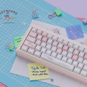 May include: A pink and white mechanical keyboard with a variety of keycap designs, including checkered and striped patterns. The keyboard sits on a blue grid-patterned surface with sticky notes and paper clips scattered around. The text "To do list" is visible on a yellow sticky note.
