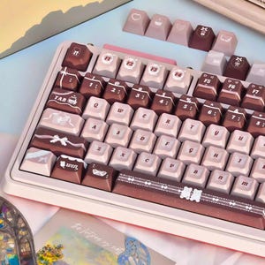 Könnte beinhalten: Eine individuelle mechanische Tastatur in Hellrosa und Braun. Die Tastenkappen sind eine Mischung aus Hellrosa und Braun mit dekorativen Symbolen und Text. Die Tastatur hat einen weißen Rahmen und ist für komfortables Tippen abgewinkelt. Ca. 30 cm breit.
