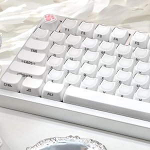 May include: A white mechanical keyboard with cat-themed keycaps. The keyboard features a pink paw print keycap and white keycaps with a cat face design. The keyboard is on a white surface.