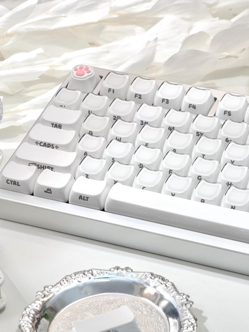 May include: A white mechanical keyboard with cat-themed keycaps. The keyboard features a pink paw print keycap and white keycaps with a cat face design. The keyboard is on a white surface.