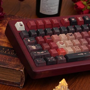 May include: A custom mechanical keyboard with a dark red wooden frame and keycaps in shades of red, black, and cream. The keycaps feature unique illustrations and text. The keyboard is angled towards the viewer, showcasing its design.