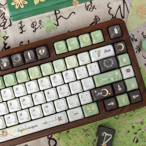 Puede incluir: Un teclado con marco de madera con teclas blancas, verdes y marrones. Las teclas presentan varios s&iacute;mbolos y dise&ntilde;os florales. La barra espaciadora tiene el texto "Euphemialynn". Elementos decorativos adicionales incluyen recortes de papel y un libro.
