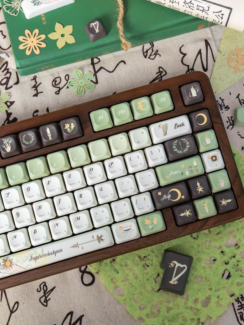 Puede incluir: Un teclado con marco de madera con teclas blancas, verdes y marrones. Las teclas presentan varios s&iacute;mbolos y dise&ntilde;os florales. La barra espaciadora tiene el texto "Euphemialynn". Elementos decorativos adicionales incluyen recortes de papel y un libro.