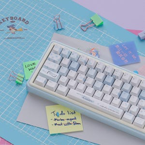 Peut inclure: Un clavier mécanique bleu clair et blanc avec des touches personnalisées. Le clavier est posé sur une surface bleue à motif quadrillé avec des post-it indiquant "To do list" et "Let it be". Des trombones décoratifs sont éparpillés.
