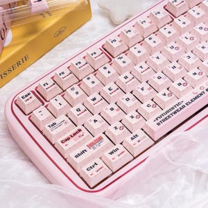Könnte beinhalten: Eine rosa mechanische Tastatur mit weißen Tastenkappen. Die Tastatur hat eine rechteckige Form mit abgerundeten Ecken und einem roten Rand. Die Tastenkappen sind mit weißen Buchstaben und Symbolen beschriftet. Der Text "<FUTURISTIC> STREETWEAR ELEMENT" ist auf der Leertaste aufgedruckt.