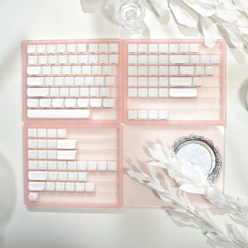 May include: A pink keyboard key organizer with white keycaps. The organizer is divided into four sections, with a small decorative mirror and white floral accents. The keyboard keys are arranged in a grid pattern.