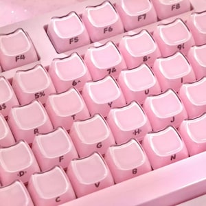 Pode incluir: Uma imagem em close-up de um teclado rosa com teclas em forma de orelhas de gato. As teclas são rosa claro com letras e símbolos brancos. O teclado tem um design moderno e minimalista, com foco na estética.