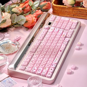 May include: A pink mechanical keyboard with custom keycaps featuring letters and cute illustrations. The keyboard sits on a pink surface, surrounded by roses and decorative items. The keyboard has a silver frame.