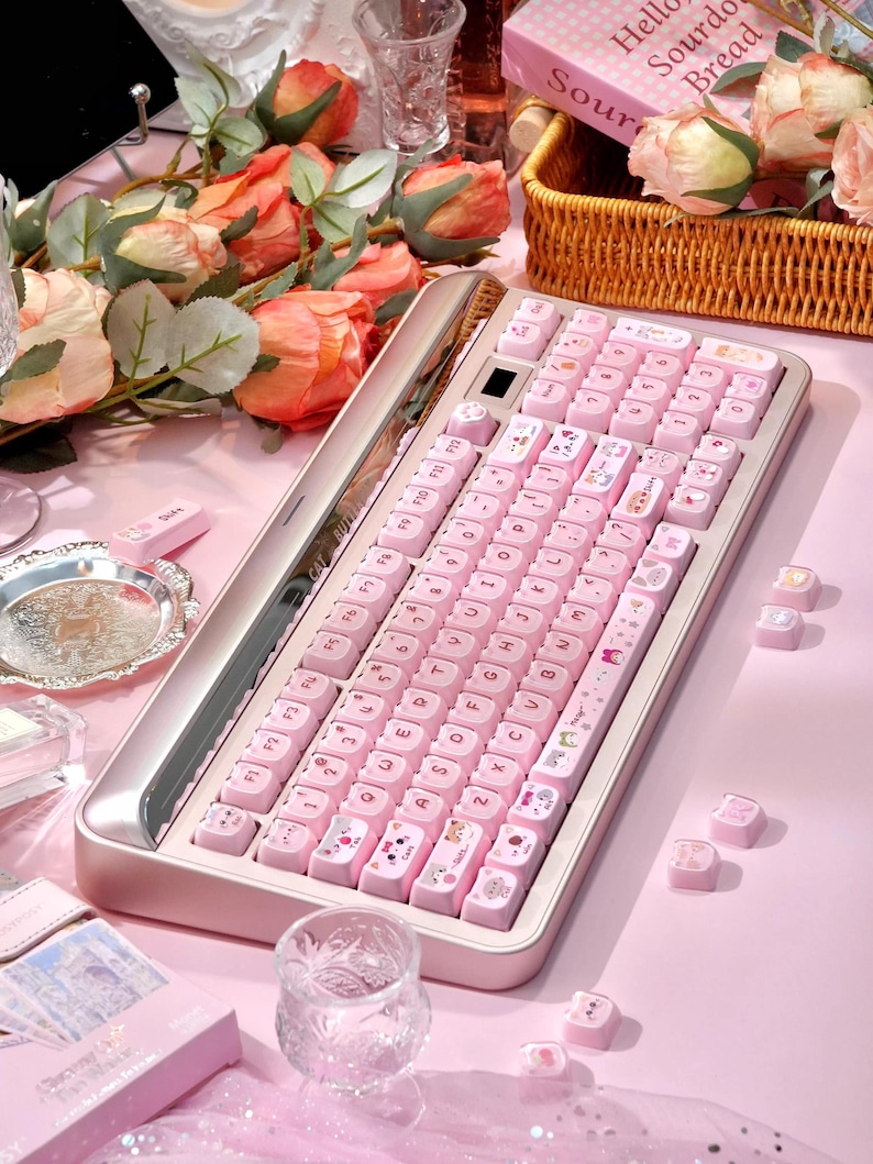 May include: A pink mechanical keyboard with custom keycaps featuring letters and cute illustrations. The keyboard sits on a pink surface, surrounded by roses and decorative items. The keyboard has a silver frame.