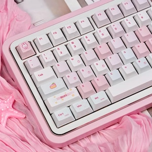 May include: A pink and white mechanical keyboard with a pastel aesthetic. The keyboard features white keycaps with pink accents and symbols, including a bunny and a cake. The keyboard is surrounded by pink fabric and a pink starfish.