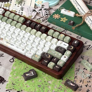 Puede incluir: Un teclado mec&aacute;nico personalizado con marco de madera y teclas en tonos blanco, verde y marr&oacute;n. Las teclas presentan varias ilustraciones, incluyendo lunas, flores y dise&ntilde;os celestiales. El teclado est&aacute; rodeado de objetos decorativos como libros y pergaminos.