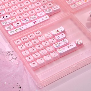 May include: A pink keyboard with white keycaps decorated with cat illustrations. The keycaps display various cat designs and symbols. The keyboard is housed in a clear pink case, with a paw-shaped keycap. The keyboard is designed for a computer.