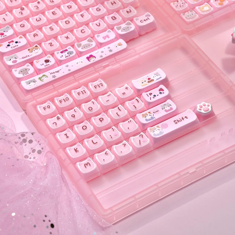 May include: A pink keyboard with white keycaps decorated with cat illustrations. The keycaps display various cat designs and symbols. The keyboard is housed in a clear pink case, with a paw-shaped keycap. The keyboard is designed for a computer.