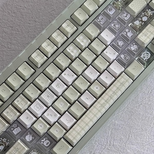 Puede incluir: Un teclado mec&aacute;nico verde claro con teclas blancas y transparentes. Algunas teclas tienen ilustraciones y patrones bonitos. El teclado tiene el texto "Telling about luck and expectations" en la parte inferior.