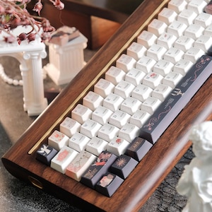 May include: A custom mechanical keyboard with a wooden frame and white keycaps. Some keycaps feature floral and artistic designs. The keyboard has a brass accent and is set against a blurred background with decorative elements.