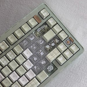 Puede incluir: Un teclado mec&aacute;nico personalizado con un marco verde claro y teclas blancas. Algunas teclas son transparentes con ilustraciones bonitas, mientras que otras tienen patrones de corazones y cuadros. El teclado tiene una tecla de acento marr&oacute;n y el texto "Telling about luck and expectations".